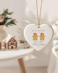 Heart-shaped ceramic Christmas ornament hanging on a gold cord, featuring two gingerbread women with bows and the text ‘Our First Christmas As Mrs & Mrs - Ella & Sophie 2025’. Styled in a warm, softly lit Christmas setting with gingerbread houses and seasonal decor in the background.