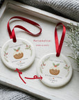 Two personalised ‘With Our Little Pudding’ first Christmas ornaments showing name and year options, displayed on a white wooden tray with festive greenery and a red ribbon.