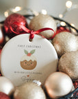Personalised ‘With Our Little Pudding’ first Christmas ornament with smiling pudding illustration, displayed among festive gold and red baubles with warm fairy lights.