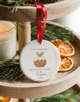 Personalised ‘First Christmas - With Our Little Pudding’ ornament with pudding illustration and holly details, hanging from a red ribbon on a wooden tiered tray beside a candle and dried orange slices.