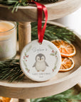 Personalised penguin-themed ceramic Christmas ornament hanging on a wooden tiered tray, surrounded by dried orange slices and spruce foliage. The design includes a baby penguin framed by holly and pine, with “Sophie’s First Christmas 2025” printed beneath. Finished with a red satin ribbon