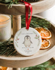Personalised ceramic baby’s first Christmas penguin ornament hanging from a red ribbon on a wooden tiered stand. The bauble features holly and pine illustrations and the text “First Christmas - With Our Little One”. Styled with pine sprigs, dried orange slices, and a lit Christmas candle