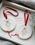 Two personalised ceramic penguin Christmas ornaments displayed on a white wooden tray beside winter greenery, both showing the phrase ‘First Christmas - With Our Little One’ with editable names and dates, highlighted with red arrows