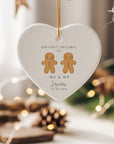 Personalised Mr & Mr gingerbread couple heart ornament with “Our First Christmas As Mr & Mr Lawson 01.09.2025” printed beneath two gingerbread men. The ceramic bauble hangs from a gold cord against a cosy Christmas scene with blurred fairy lights and natural wooden décor