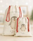 Luxury personalised Christmas sacks featuring hand-illustrated penguin designs, shown in a softly lit festive room with blurred twinkling lights. One large Santa sack and one small gift sack stand side by side, both made from premium linen-look fabric with red drawcord ties. A warm, nostalgic Christmas aesthetic.