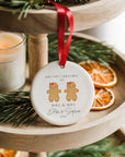 A round ceramic Christmas ornament featuring two gingerbread women with red bows and a heart above them, personalised with “Our First Christmas as Mrs & Mrs” and the names Ella and Sophie with the year 2025. The ornament hangs from a red ribbon on a wooden tiered tray decorated with pine sprigs, dried orange slices, and a softly glowing candle for a cosy festive atmosphere