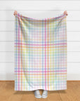 Length of large scale pastel rainbow gingham fabric held against a white shiplap wall, showing the soft pink, blue, lilac, mint and peach check.