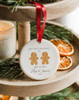 A round ceramic Christmas ornament featuring two gingerbread men with a small red heart above them, personalised with “Our First Christmas as Mr & Mr” and the names Alex and James with the year 2025. The ornament hangs from a red ribbon on a wooden tiered tray decorated with pine branches, dried orange slices, and a softly glowing candle for a cosy festive atmosphere