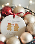 A personalised ceramic ornament showing a gingerbread couple design with red bows and a heart, printed with the text “Our First Christmas as Fiancés” and the names Ella and Sophie. The ornament is nestled in a basket filled with glittery gold and red baubles and warm fairy lights for a magical Christmas atmosphere.