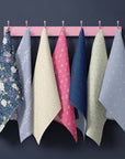 Spring Gardens At Night blue floral tea towel collection hanging on pink hooks against a deep charcoal wall, featuring navy, lavender, butter yellow, coral pink and soft sage cottagecore patterns.