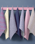 Seven floral tea towels hanging on blush pink hooks against a soft blue wall, featuring navy, powder blue, butter yellow and soft pink prints including ditsy florals, delicate stripes and scattered blossoms in a spring cottage palette.