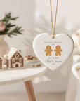 Heart-shaped ceramic Christmas ornament featuring two gingerbread men and the text “Our First Christmas As Mr & Mr Alex & James 2025.” The bauble hangs from a gold cord in a warm, softly lit festive setting with gingerbread houses and neutral Christmas décor in the background