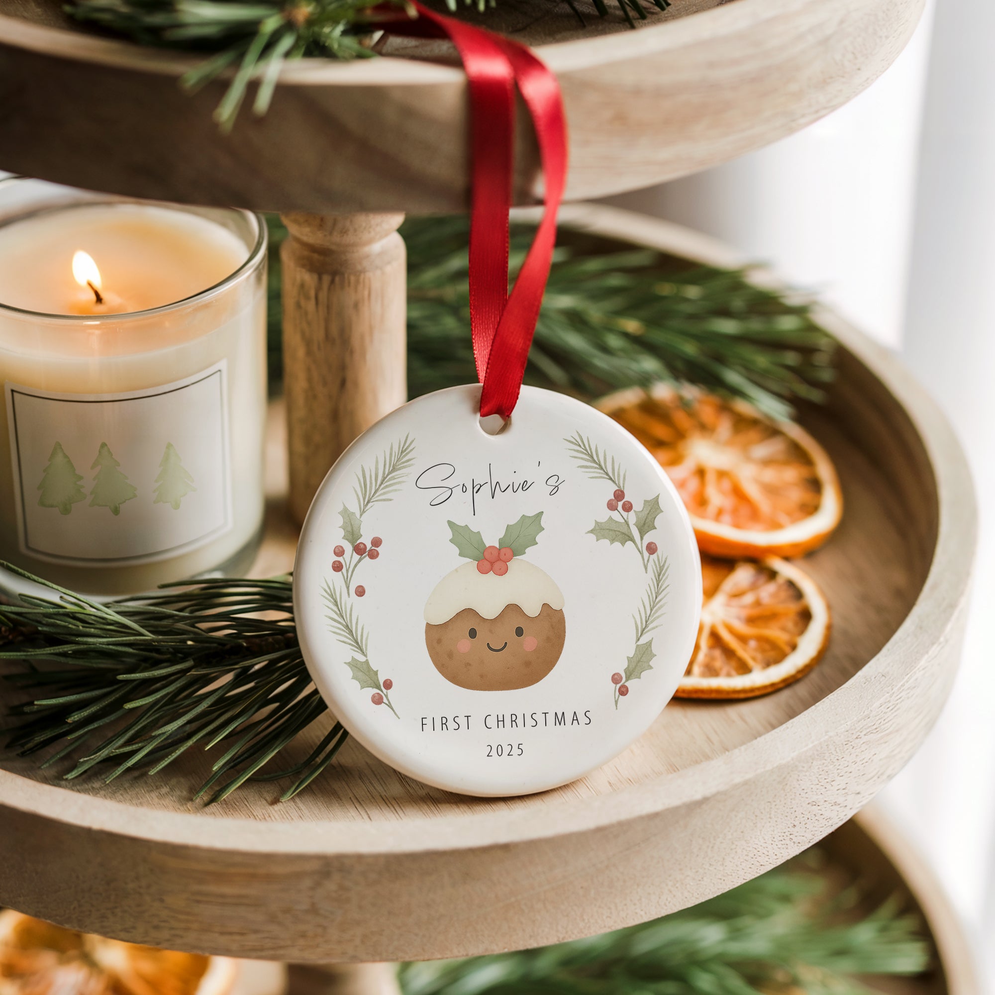 Personalised baby’s first Christmas pudding ornament with holly and pine wreath design, hanging from a red ribbon on a wooden tiered tray beside a candle and dried orange slices.