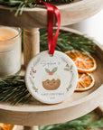 Personalised baby’s first Christmas pudding ornament with holly and pine wreath design, hanging from a red ribbon on a wooden tiered tray beside a candle and dried orange slices.