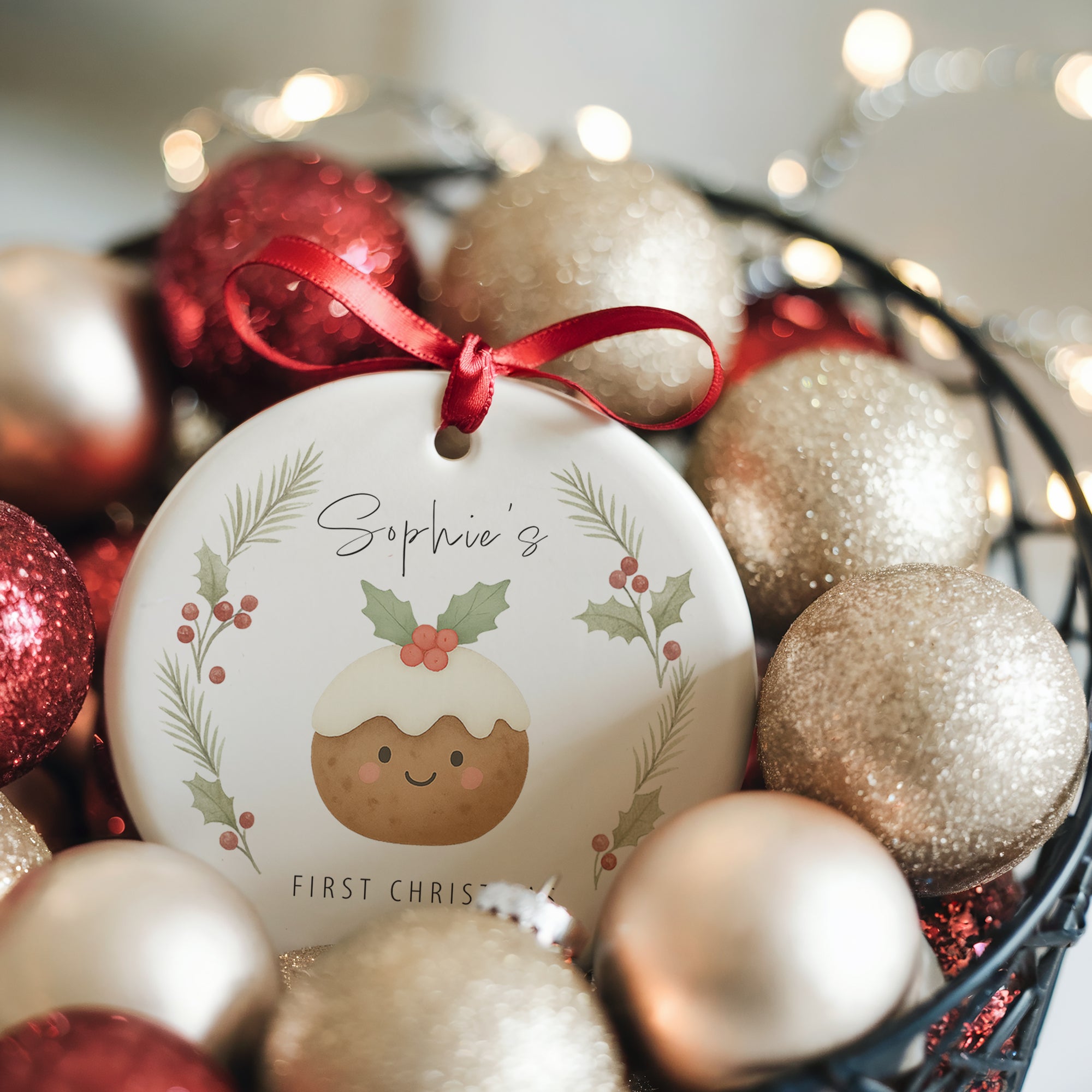 Personalised Christmas pudding ornament with festive holly and pine wreath design, displayed in a bowl of red and gold baubles with warm fairy lights
