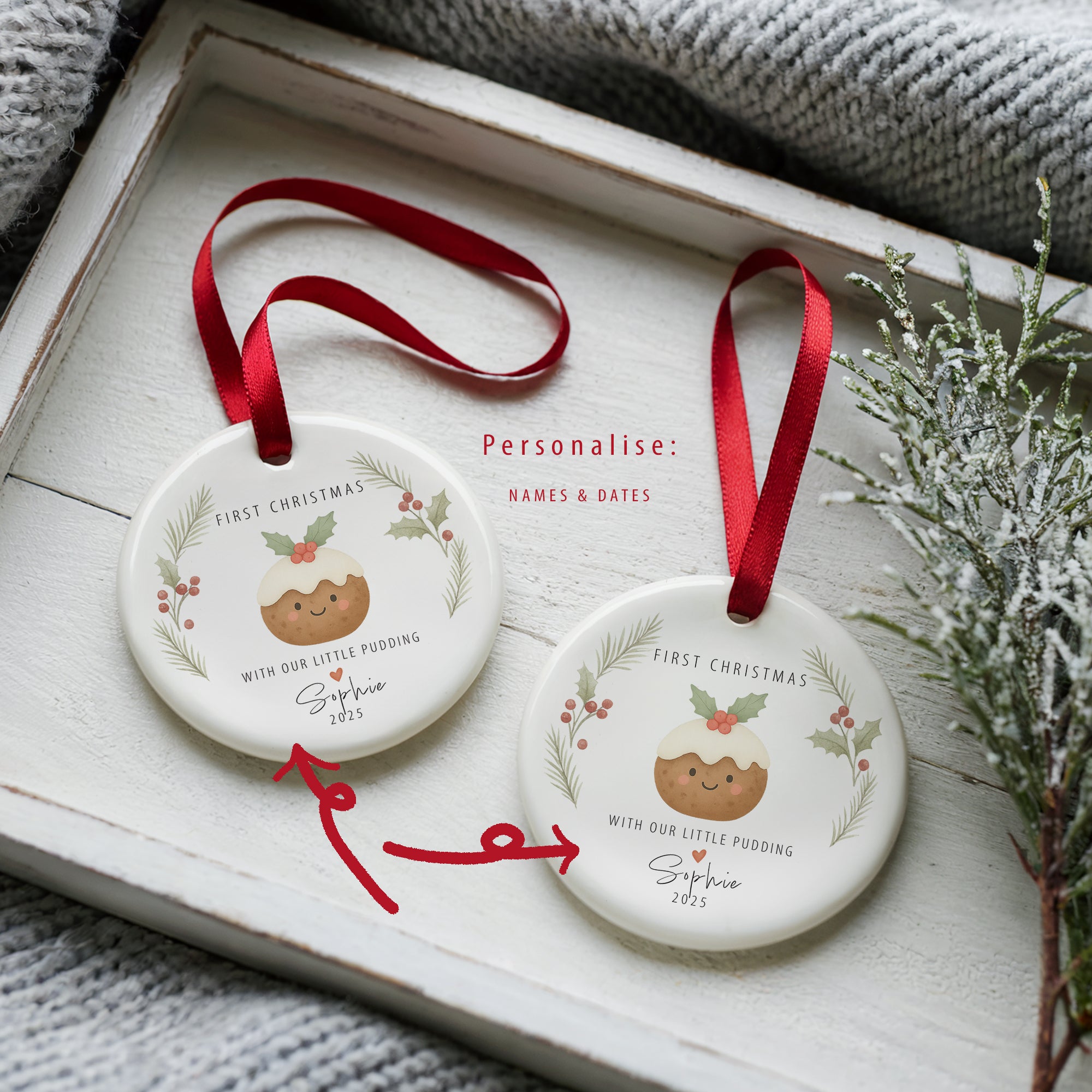 Two personalised ‘With Our Little Pudding’ first Christmas ornaments showing name and year options, displayed on a white wooden tray with festive greenery and a red ribbon.