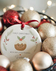 Personalised ‘With Our Little Pudding’ first Christmas ornament with holly and pine wreath design, displayed in a bowl of red and gold baubles with warm fairy lights