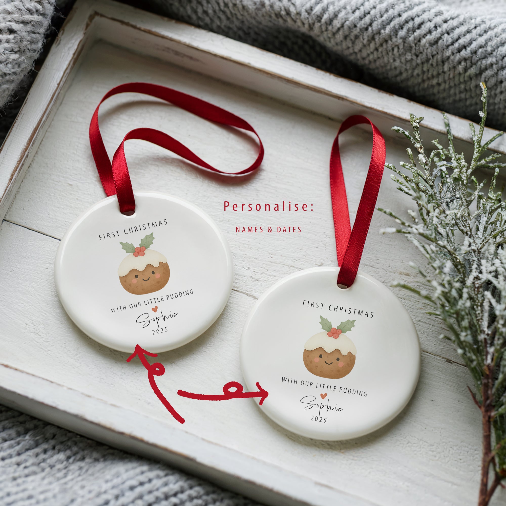 Two personalised ‘With Our Little Pudding’ first Christmas ornaments showing custom name and year options, displayed on a white wooden tray with festive greenery and red ribbons.