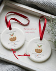 Two personalised ‘With Our Little Pudding’ first Christmas ornaments showing custom name and year options, displayed on a white wooden tray with festive greenery and red ribbons.