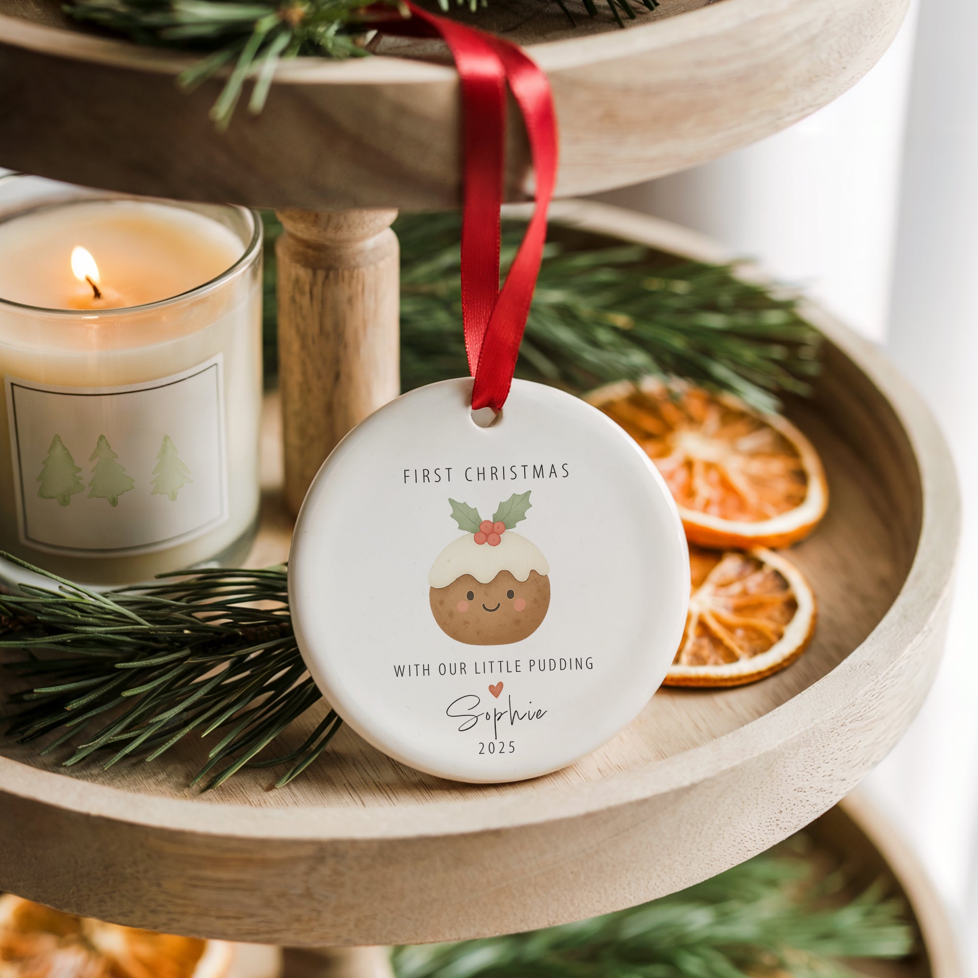 Personalised ‘First Christmas - With Our Little Pudding’ ornament with pudding illustration and holly details, hanging from a red ribbon on a wooden tiered tray beside a candle and dried orange slices.