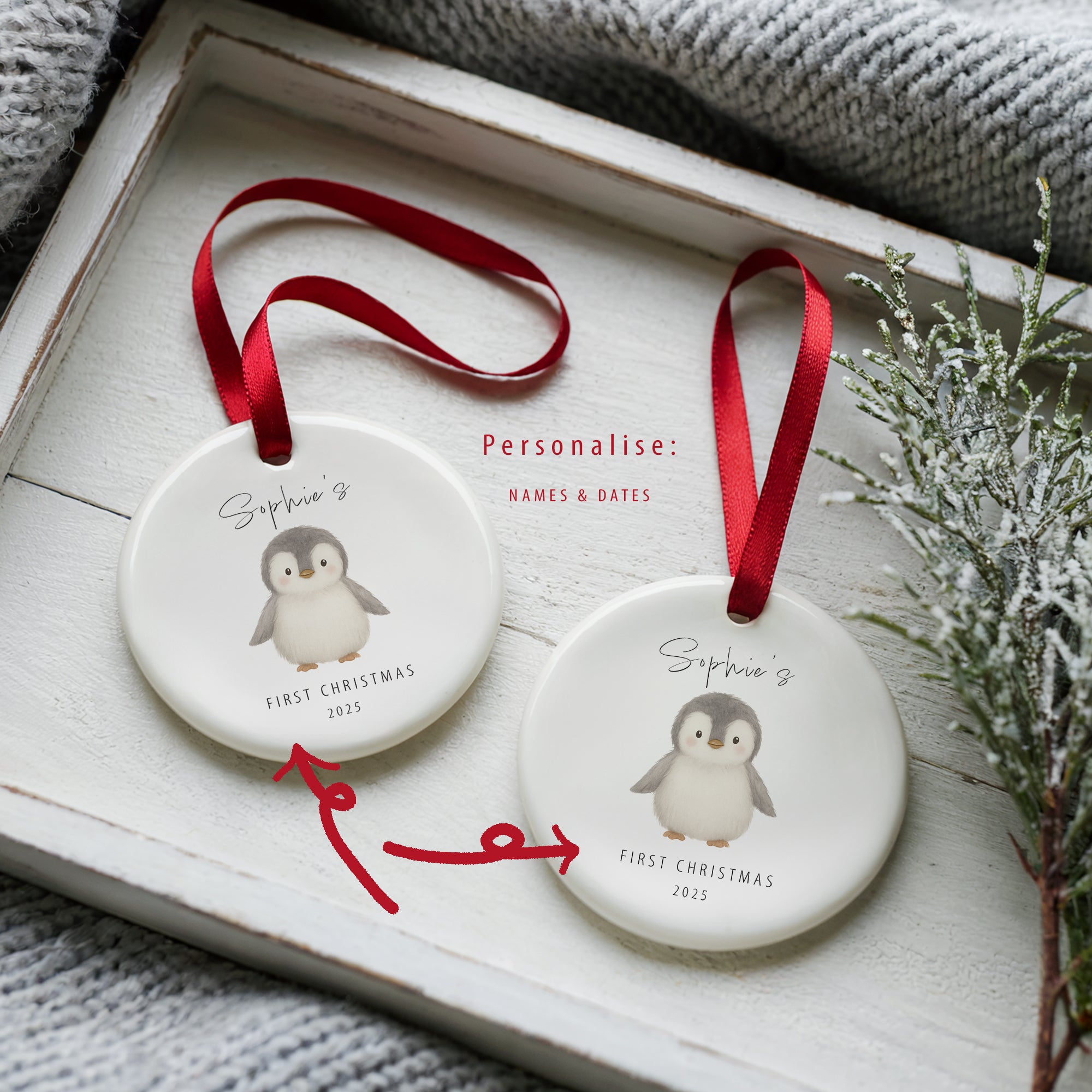Two personalised baby’s first Christmas penguin ornaments shown side by side in a white wooden tray, each printed with name and year. Red arrows highlight the customisable areas, with a festive sprig and soft blanket in the background