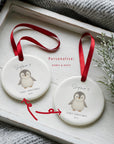 Two personalised baby’s first Christmas penguin ornaments shown side by side in a white wooden tray, each printed with name and year. Red arrows highlight the customisable areas, with a festive sprig and soft blanket in the background
