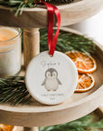 Baby’s first Christmas ceramic ornament with a sweet penguin illustration and the name ‘Sophie’, hanging from a red ribbon on a wooden tiered display stand with dried oranges, pine greenery and a lit candle beside it.