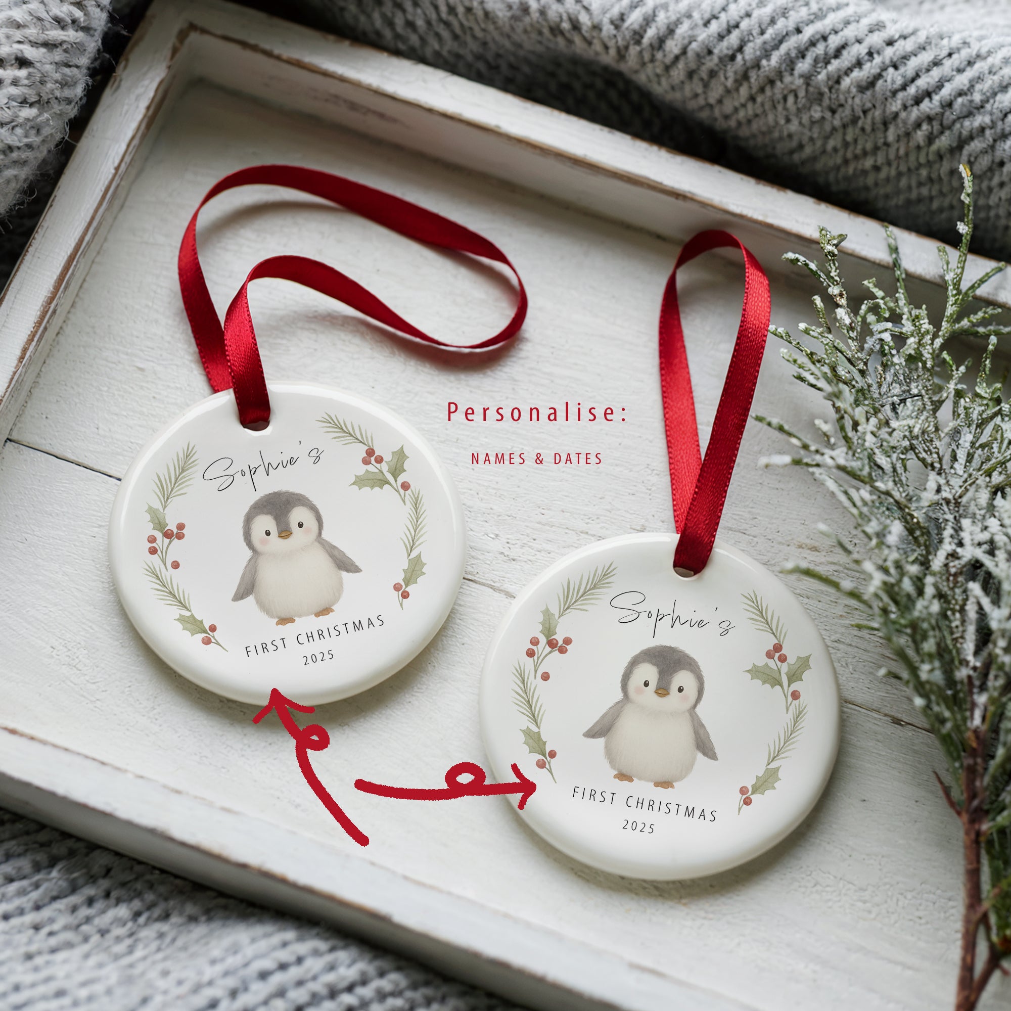 Two round ceramic penguin Christmas ornaments displayed in a white wooden tray beside winter greenery. Each features a penguin illustration framed with holly and pine, with space for a personalised name and date. Red arrows highlight the customisation options for names and dates. Both hang from red satin ribbon