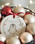 Image showing a personalised round ceramic Christmas ornament featuring a baby penguin illustration, surrounded by soft holly and pine sprigs. The ornament hangs from a red satin ribbon among gold and red baubles with warm fairy lights in the background. The name “Sophie’s” appears above the penguin with “First Christmas 2025
