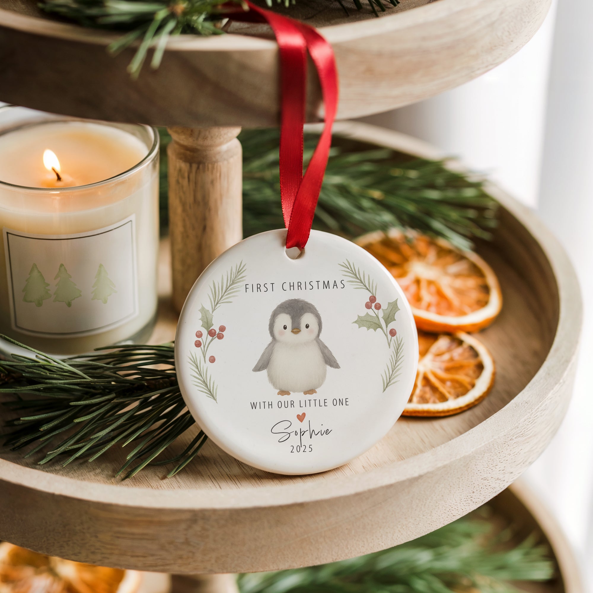 Personalised ceramic baby’s first Christmas penguin ornament hanging from a red ribbon on a wooden tiered stand. The bauble features holly and pine illustrations and the text “First Christmas - With Our Little One”. Styled with pine sprigs, dried orange slices, and a lit Christmas candle