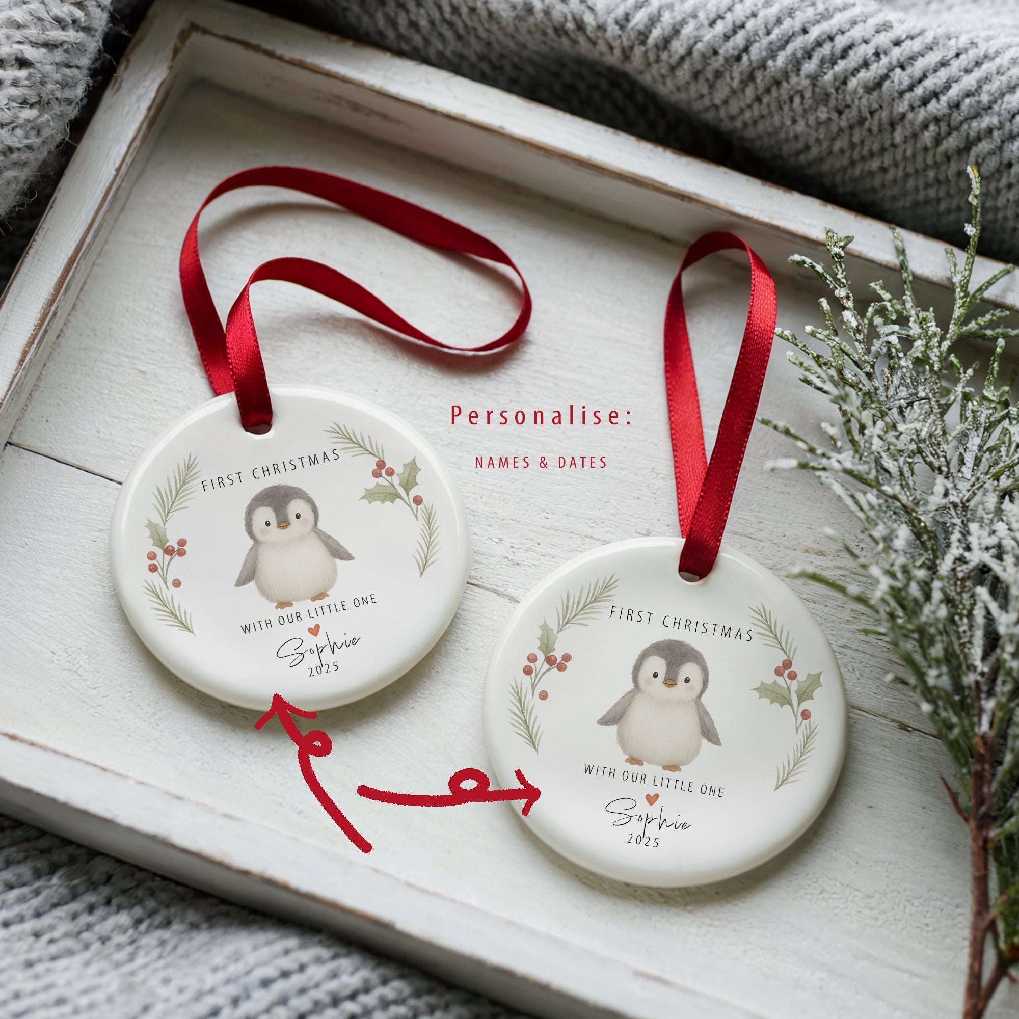 Two personalised ceramic ‘First Christmas – With Our Little One’ penguin ornaments displayed on a white wooden tray with red satin ribbons. One ornament is annotated with arrows showing where names and dates can be customised, styled beside a soft grey blanket and festive greenery