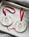Two personalised ceramic ‘First Christmas – With Our Little One’ penguin ornaments displayed on a white wooden tray with red satin ribbons. One ornament is annotated with arrows showing where names and dates can be customised, styled beside a soft grey blanket and festive greenery
