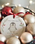 Personalised ceramic baby’s first Christmas ornament featuring a cute baby penguin framed by holly and pine illustrations. The decoration hangs from a red satin ribbon and sits nestled among gold and red baubles with warm fairy lights in the background