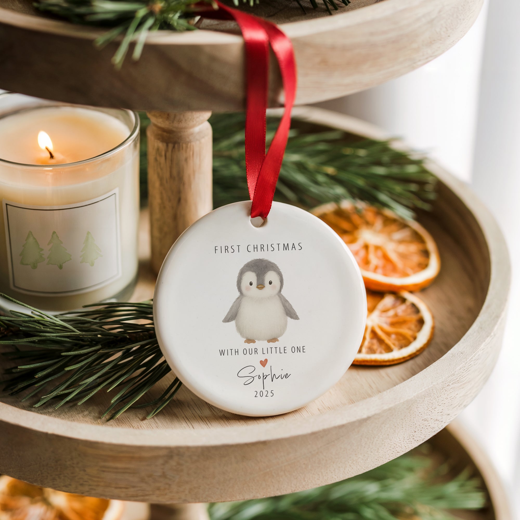 Personalised ceramic penguin ‘First Christmas - With Our Little One’ ornament hanging from a red ribbon on a wooden tiered tray, surrounded by pine sprigs and dried orange slices for a cosy festive scene