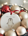 Ceramic ‘First Christmas – With Our Little One’ penguin ornament nestled in a basket of gold and red baubles, personalised with baby’s name and year, hanging from a red satin ribbon with warm fairy lights in the background.