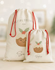 Luxury personalised Christmas sacks featuring hand-illustrated Christmas pudding designs, displayed in a softly lit festive room with blurred warm fairy lights. One large Santa sack and one smaller gift sack stand together, both made from thick linen-look fabric with bright red drawcord ties. A warm, nostalgic holiday atmosphere