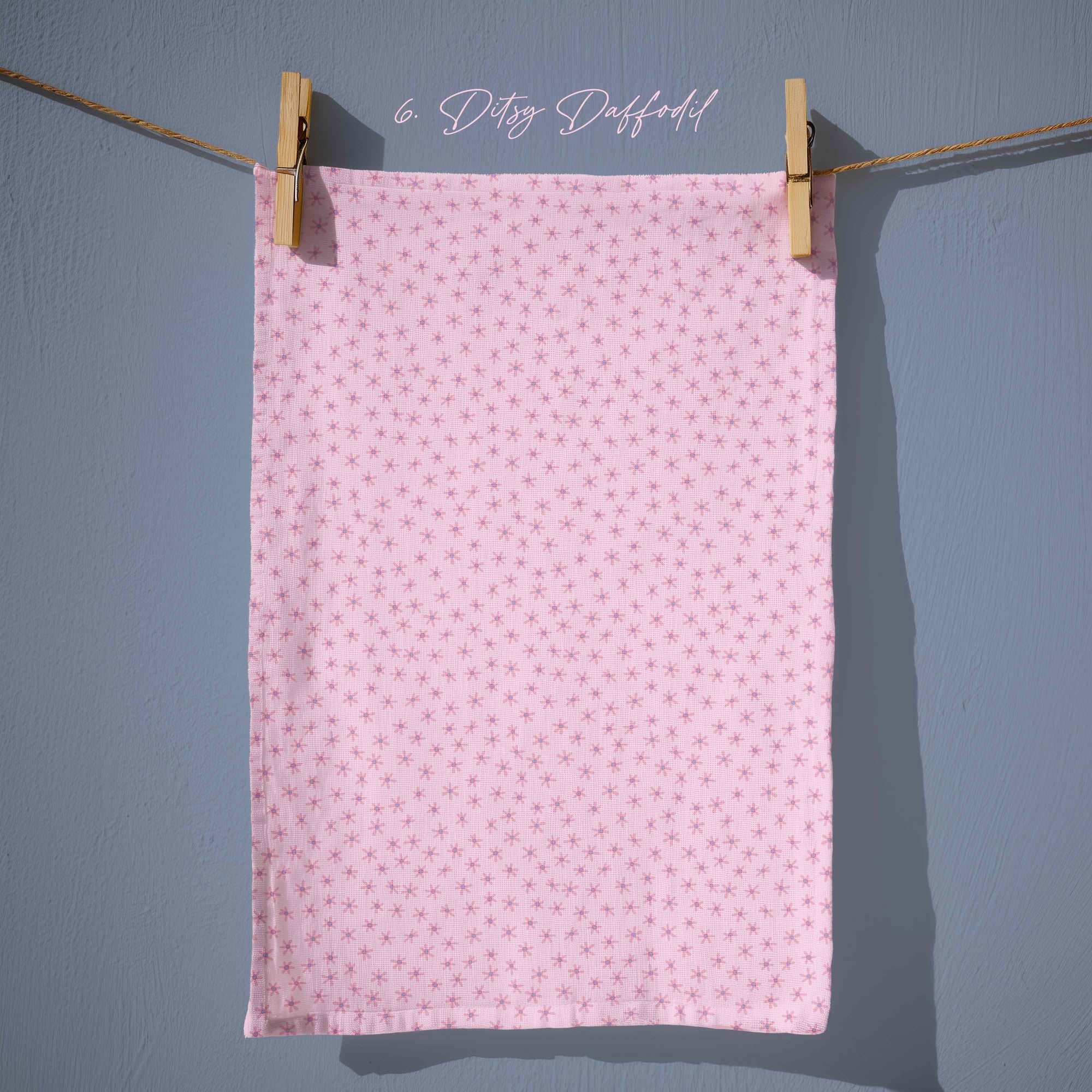 Blush pink tea towel covered in tiny scattered floral stars in lilac and rose tones, creating a sweet, ditsy spring pattern with vintage charm.