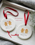 Two round ceramic gingerbread couple ornaments with red ribbons, showing the “Engaged” and “Fiancés” wording options personalised with names and dates. The ornaments are styled on a white wooden tray with winter foliage and a soft knitted blanket, highlighting the customisation options.