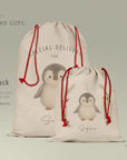 Side-by-side visual comparison of the large Santa Sack and small Gift Sack, displayed on a sage background. Both sacks feature the illustrated penguin design, thick linen-look fabric and red cords. Includes text showing the two available sizes, approximate measurements, and suggested uses such as Santa deliveries, toy sacks, reusable gift bags.
