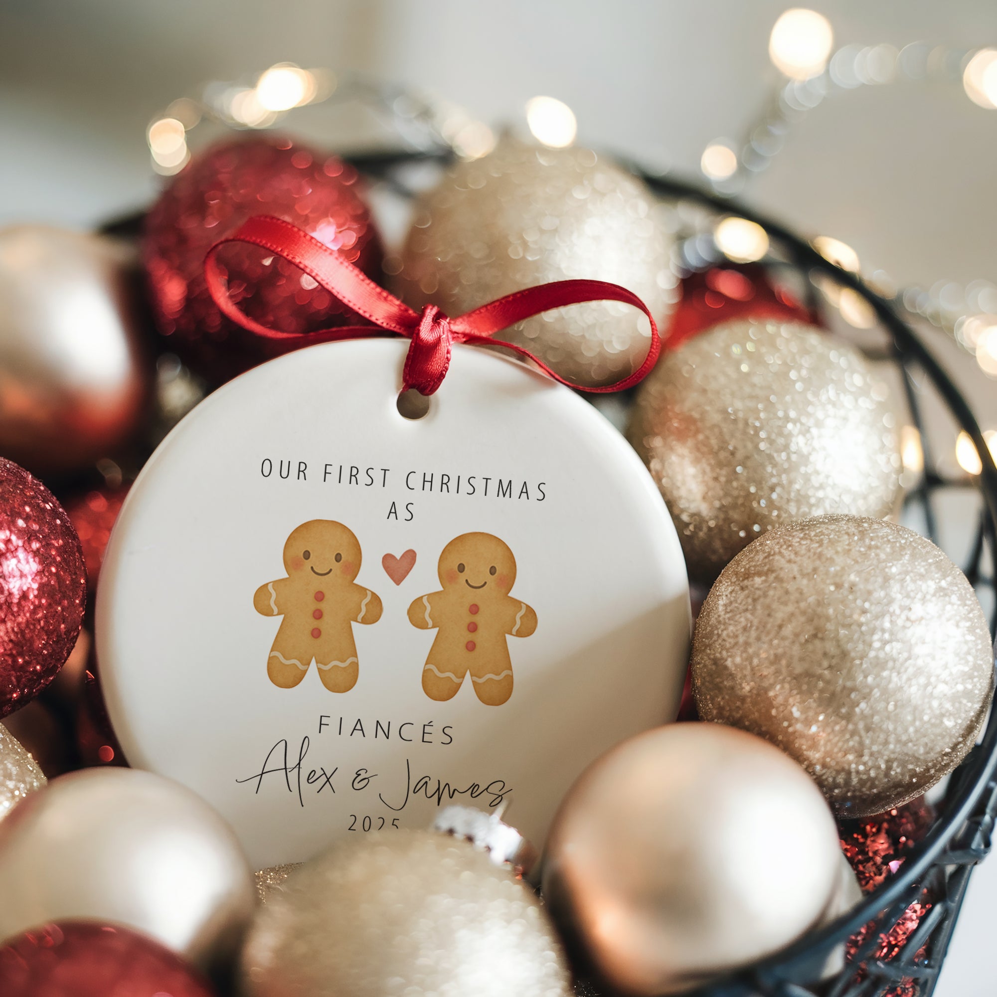 Two personalised gingerbread couple ceramic ornaments showing “Engaged” and “Fiancés” versions with red ribbons, styled on a white wooden tray with winter greenery