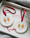 Two round ceramic gingerbread couple ornaments displaying the “Mr & Mr” personalisation options: one using first names with the year, and one showing the married surname with the full wedding date. Both ornaments hang from red ribbons and are styled on a white wooden tray with winter greenery and a cosy knitted blanket to highlight the customisation choices