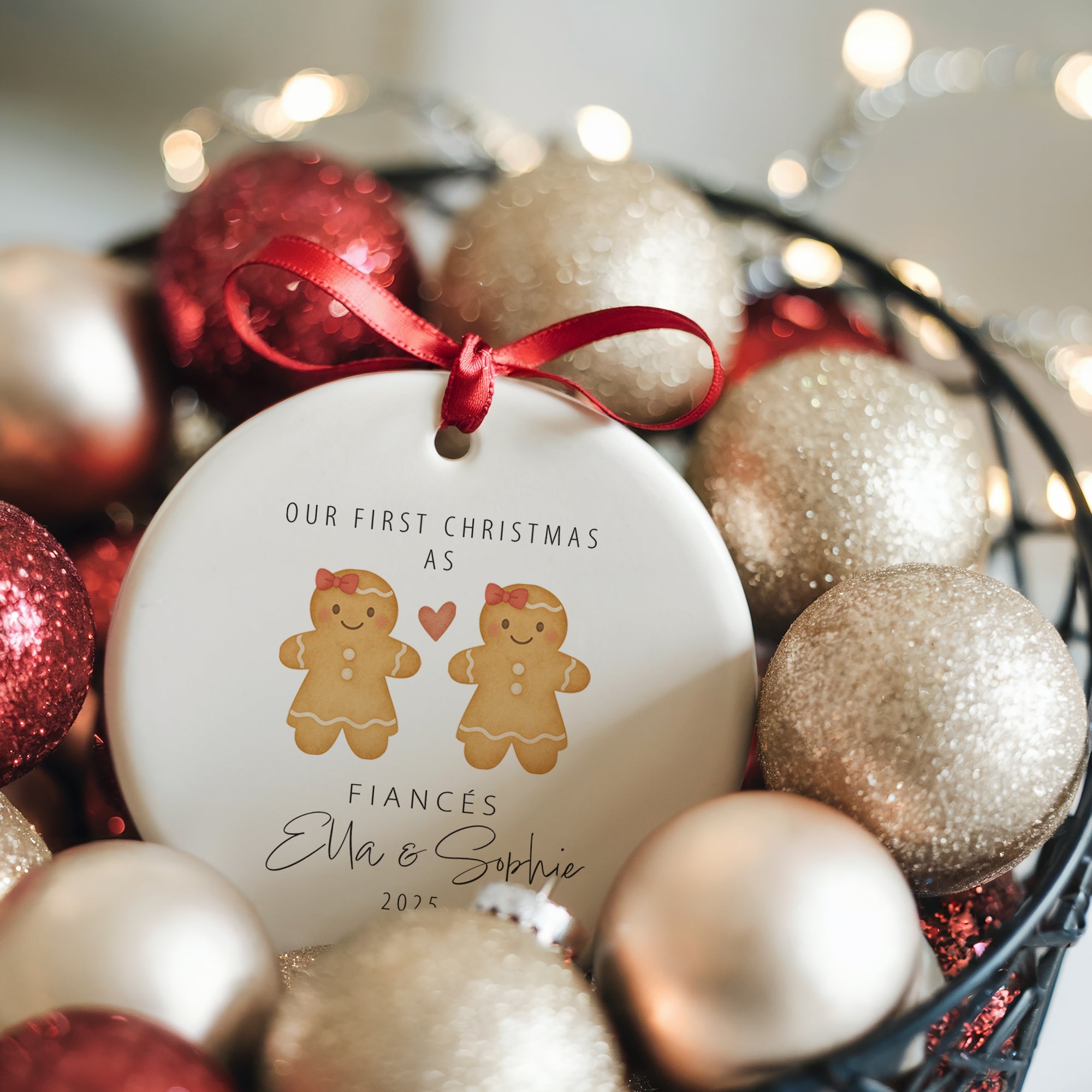 A personalised ceramic ornament showing a gingerbread couple design with red bows and a heart, printed with the text “Our First Christmas as Fiancés” and the names Ella and Sophie. The ornament is nestled in a basket filled with glittery gold and red baubles and warm fairy lights for a magical Christmas atmosphere.