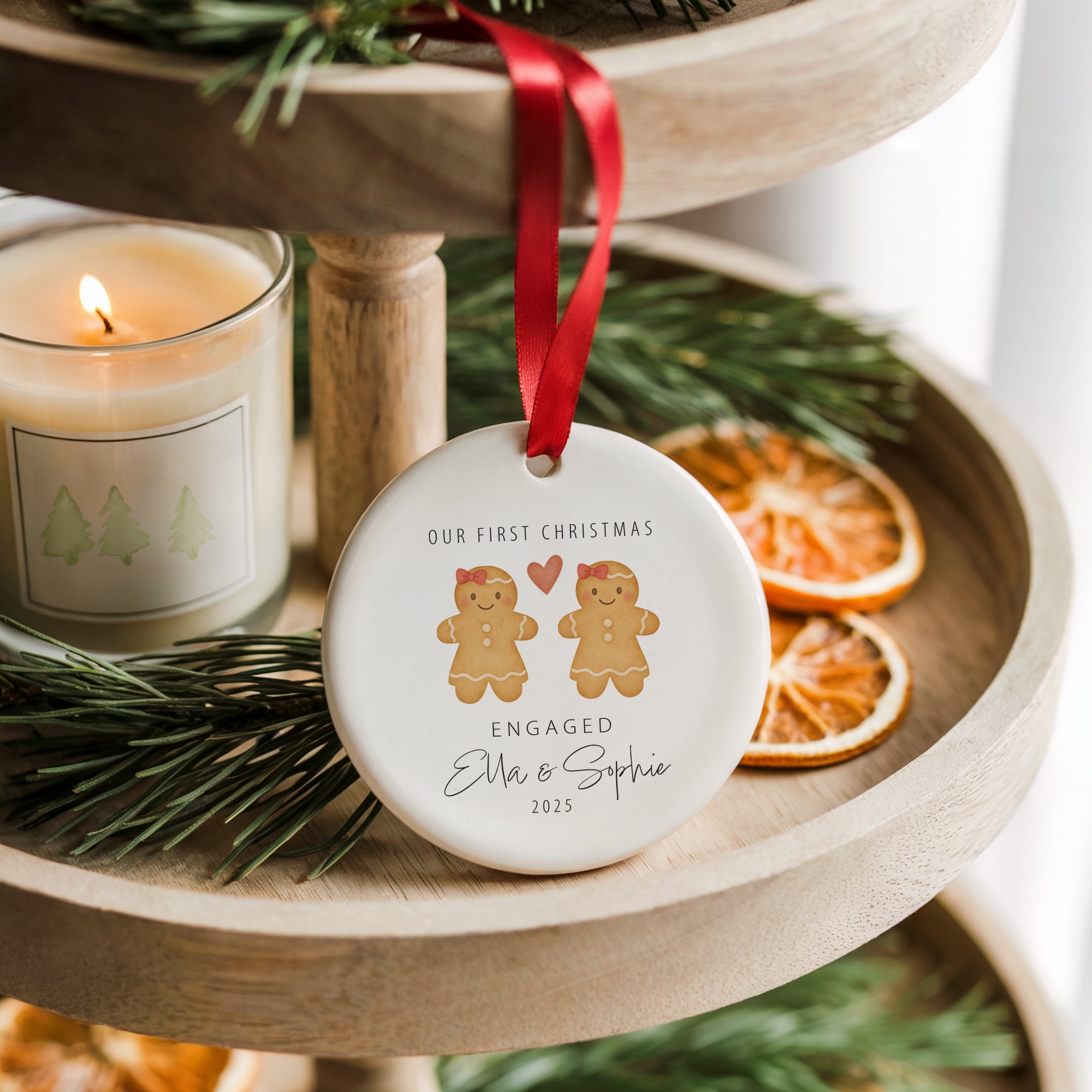 A round ceramic Christmas ornament featuring two gingerbread women with red bows and a small red heart above them, personalised with the names Ella and Sophie and the word “Engaged.” The ornament hangs from a red ribbon on a wooden tiered tray decorated with pine branches, dried orange slices, and a softly glowing candle, creating a cosy festive scene