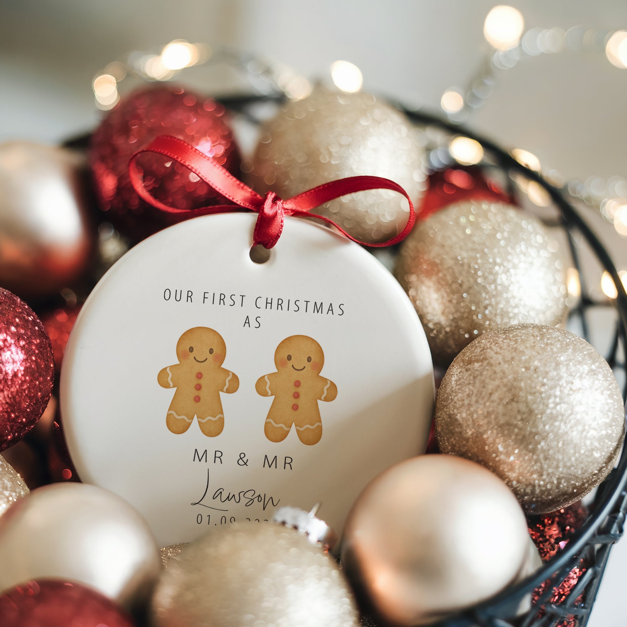 A personalised ceramic ornament showing two gingerbread men with a red heart and the text “Our First Christmas as Mr & Mr” along with the surname Lawson and wedding date. The ornament is nestled in a basket filled with glittery red and gold baubles and warm fairy lights, creating a magical Christmas scene