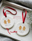 Two round ceramic gingerbread couple ornaments showing the personalisation options: one with first names and year, and one with the married surname and full wedding date. Both hang from red ribbons and are styled on a white wooden tray with winter greenery and a soft knitted blanket, highlighting the customisable details