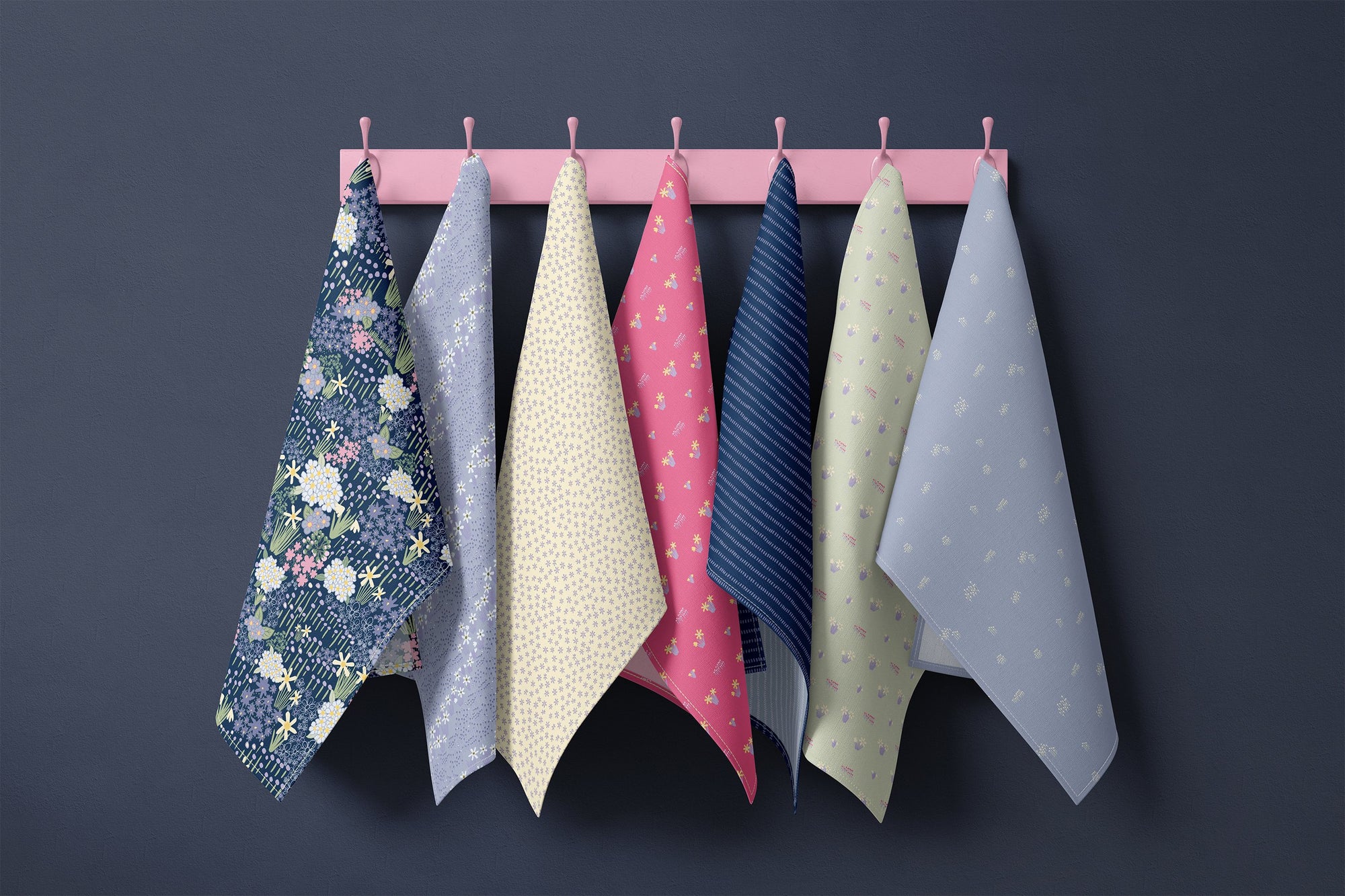 Spring Gardens At Night blue floral tea towel collection hanging on pink hooks against a deep charcoal wall, featuring navy, lavender, butter yellow, coral pink and soft sage cottagecore patterns.