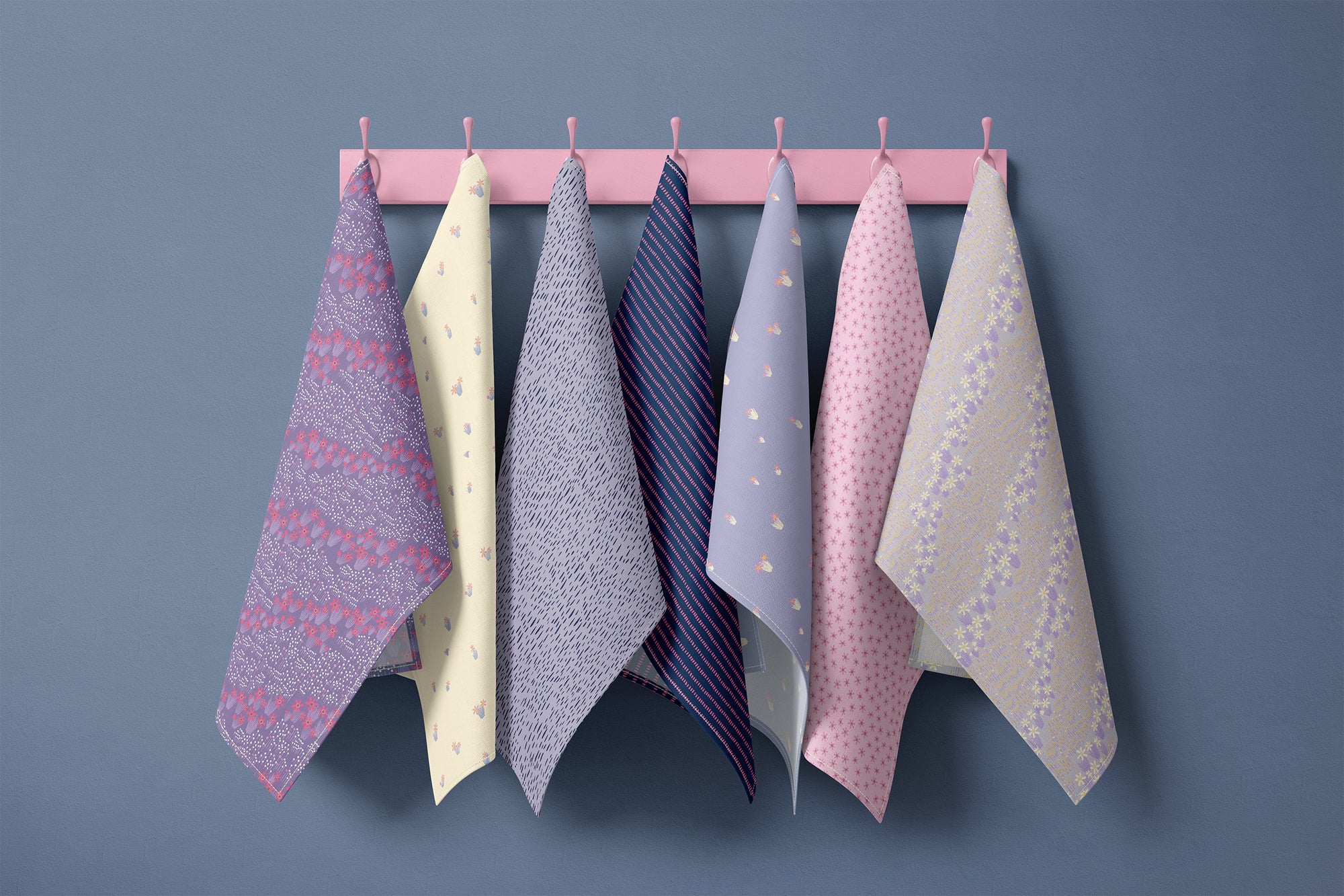 Seven floral tea towels hanging on blush pink hooks against a soft blue wall, featuring navy, powder blue, butter yellow and soft pink prints including ditsy florals, delicate stripes and scattered blossoms in a spring cottage palette.