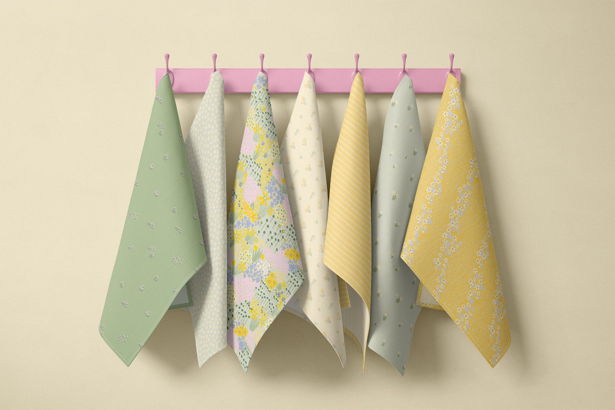 Pastel spring floral tea towel collection hanging on pink wall hooks featuring sage green, soft yellow and cottage garden prints in daffodil, blossom and stripe patterns for a country kitchen