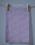 Blue tea towel with flowing trails of small pink blossoms and scattered white crocus speckles, creating a soft dusk-inspired floral pattern with gentle movement and layered texture.